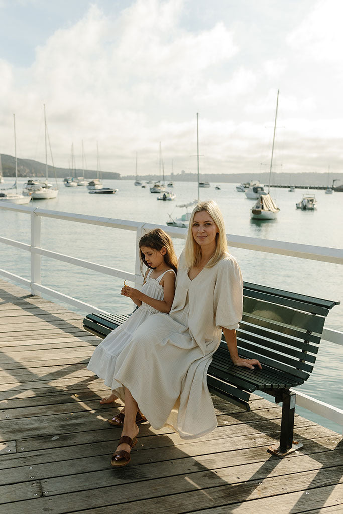 blonde haired woman and dark haired girl child sitting on a park bench with a harbour in the background